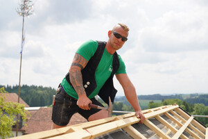 Das Bild zeigt einen Zimmerer bei seiner Arbeit. Er kniet auf einem Dachfirst, hält einen Hammer in der Hand und befestigt Dachlatten.