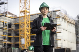 Das Bild zeigt eine junge Frau mit Bauhelm und Bau-Arbeitskleidung. Sie hält ein Tablett in der Hand und schaut selbstbewusst in die Kamera. Im Hintergrund ist ein Rohbau und ein Kran zu sehen.