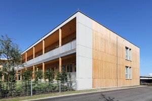 Das Bild zeigt ein 2-geschossiges Gebäude aus Holz. Die Vorderfront ist mit einem Vorbau ausgestattet, der im Erdgeschoss wie Arkaden wirkt und im ersten OG zur Außenterrasse wird.
