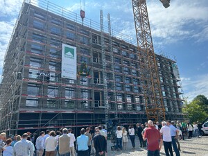 Das Foto zeigt ein Richtfest. Ein großer Kranz wird gerade vor dem fertigen 5-stöckigen Rohbau hochgelassen. Gäste stehen vor dem Bau und schauen nach oben.
